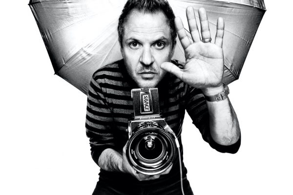 black and white portrait of photographer Platon, holding up one hand with a camera laying on the other, with camera lighting behind him